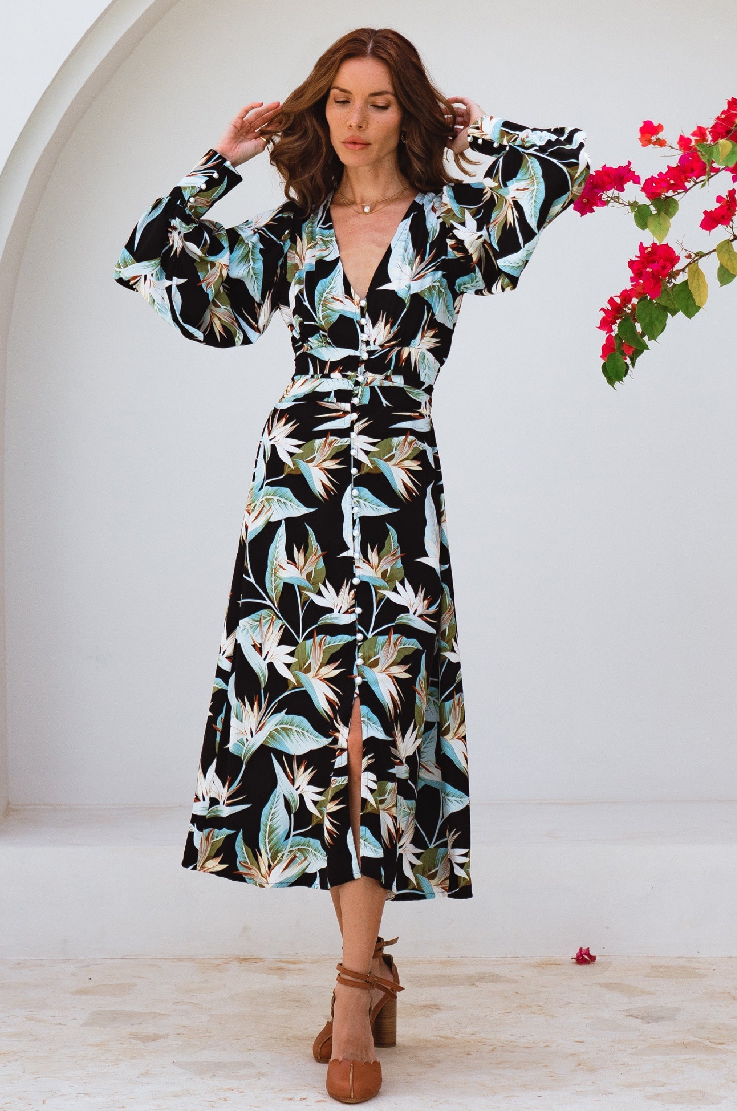 Woman wearing a floral midi dress with pearl buttons, V-neckline, balloon sleeve, and fitted detailing on the front standing with a white background and flowers in the corner, front view