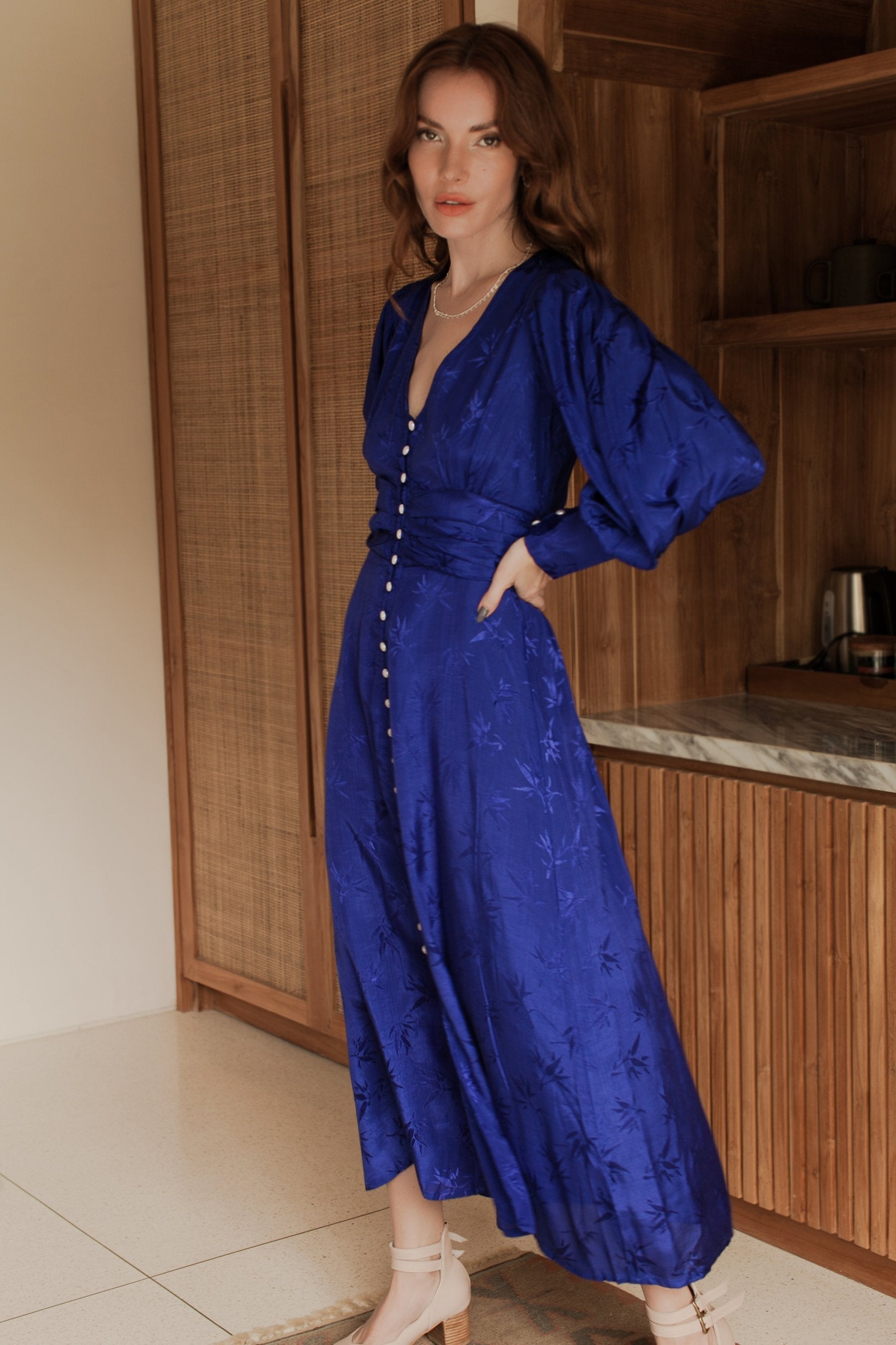 Woman wearing a blue midi dress with pearl buttons, V-neckline, balloon sleeve, and fitted detailing on the front standing in a room with wooden cabinets, 3/4 view