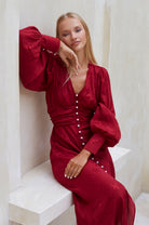 Woman in a red Woman midi dress with pearl buttons, V-neckline, balloon sleeve, and fitted detailing on the front sitting on a white bench.