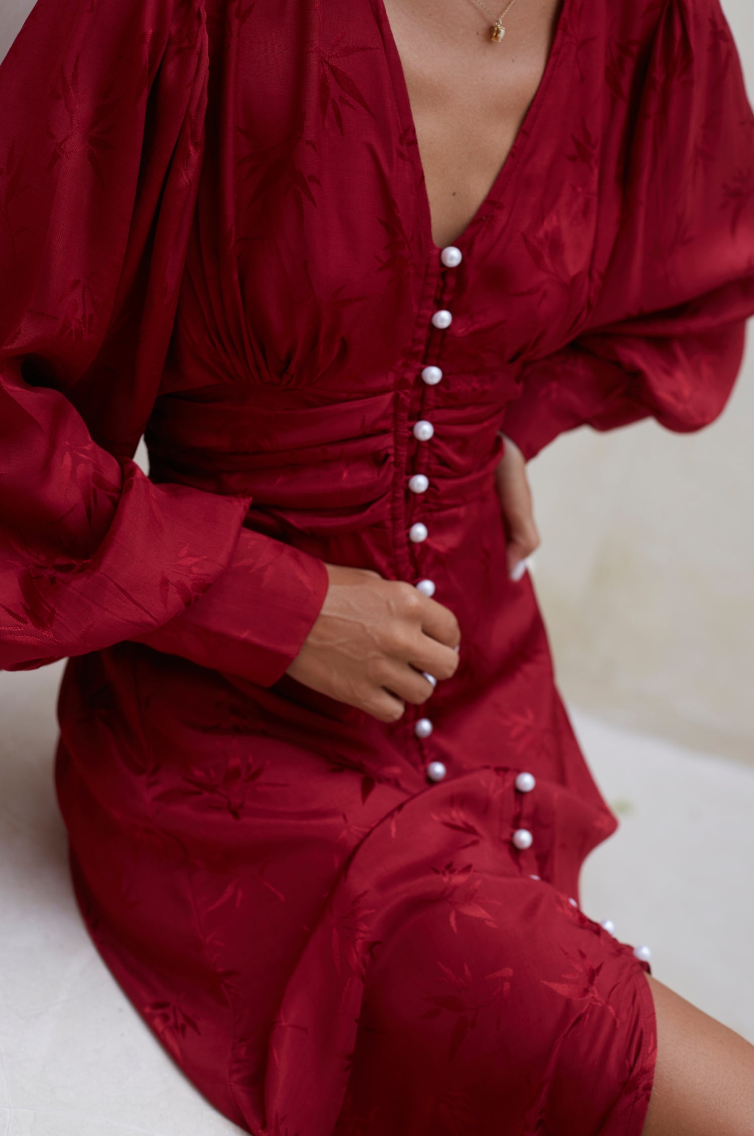 Woman in a red midi dress with pearl buttons, V-neckline, balloon sleeve, and fitted detailing on the front sitting on a white bench, closeup