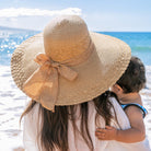 AVAH-woman wearing a large khaki Wide Brim Straw Sun Hat that features a scallop edge design and a burlap bow accent on a beach back view