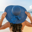 AVAH-woman wearing a large navy-blue Wide Brim Straw Sun Hat that features a scallop edge design and a burlap bow accent on a beach back view