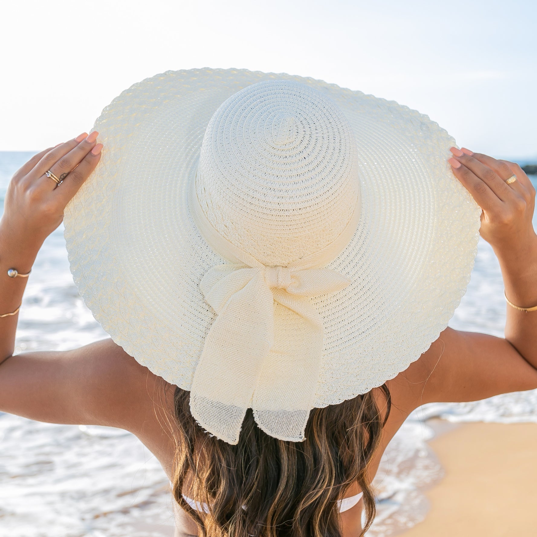 AVAH-Woman wearing a large cream Wide Brim Straw Sun Hat that features a scallop edge design and a burlap bow accent on a beach back view