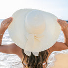 AVAH-Woman wearing a large cream Wide Brim Straw Sun Hat that features a scallop edge design and a burlap bow accent on a beach back view