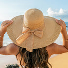 AVAH-woman wearing a large khaki Wide Brim Straw Sun Hat that features a scallop edge design and a burlap bow accent on a beach back view