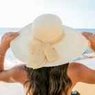 AVAH-woman wearing a large cream Wide Brim Straw Sun Hat that features a scallop edge design and a burlap bow accent on a beach back view