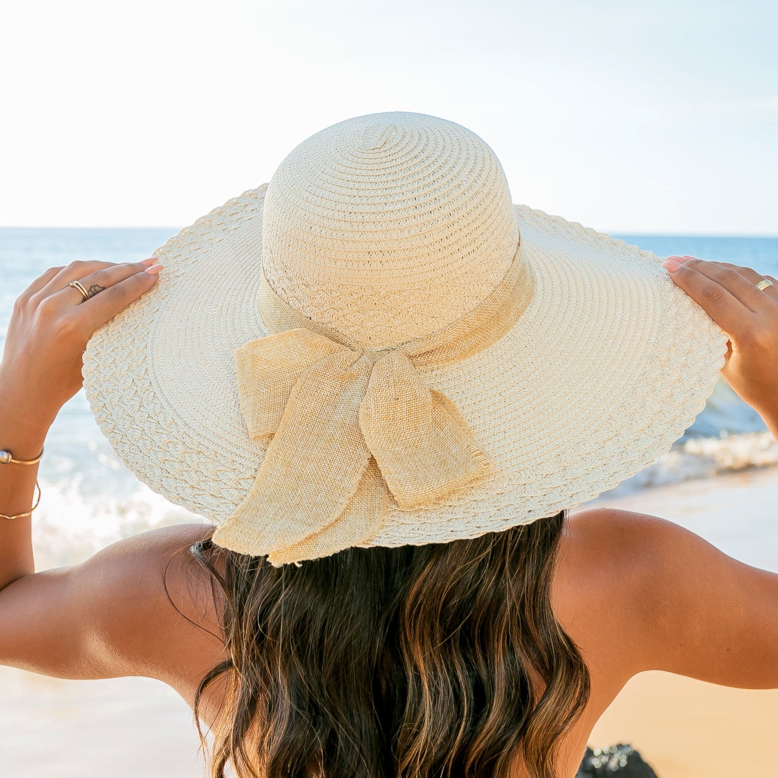 AVAH-woman wearing a large cream Wide Brim Straw Sun Hat that features a scallop edge design and a burlap bow accent on a beach back view