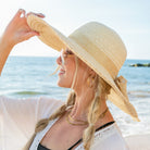 AVAH-woman wearing a large cream Wide Brim Straw Sun Hat that features a scallop edge design and a burlap bow accent on a beach side view