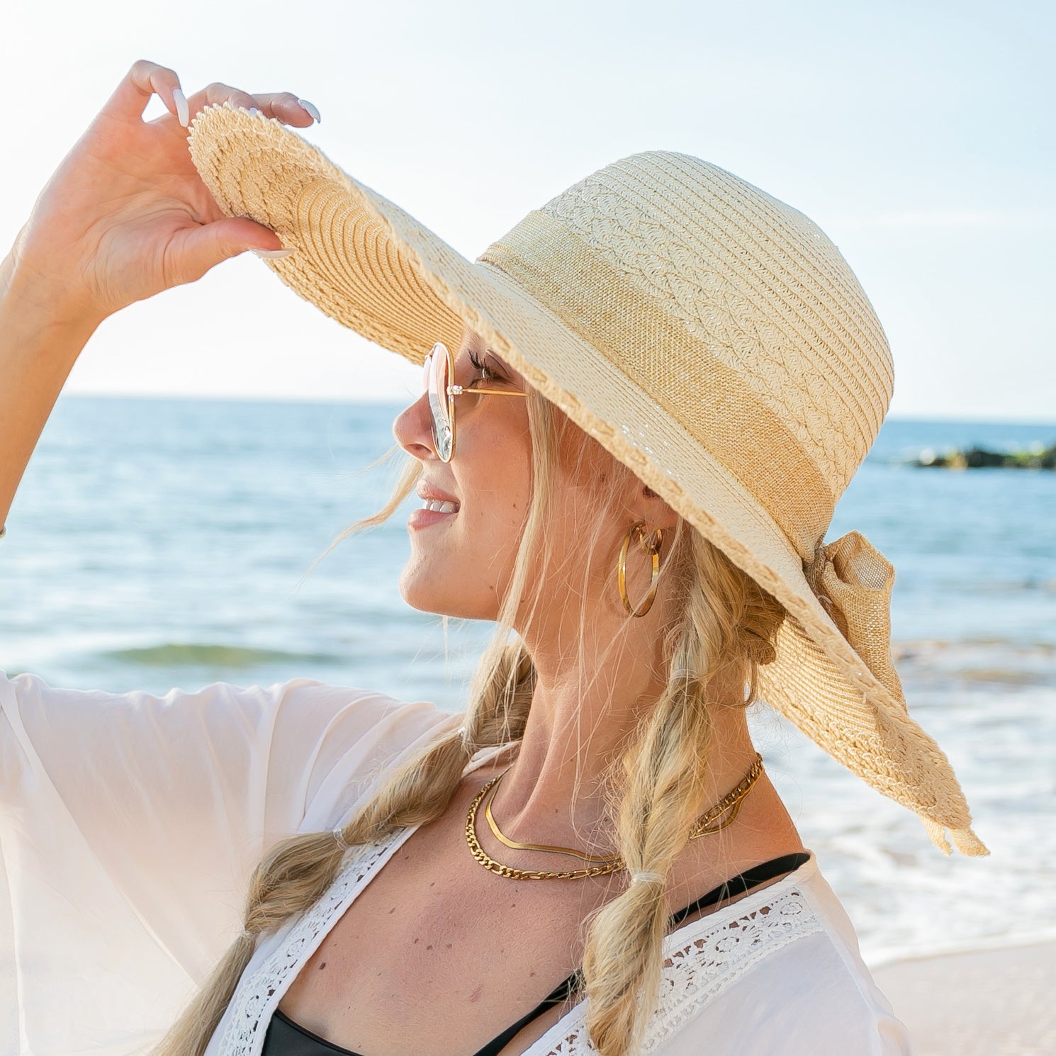 AVAH-woman wearing a large cream Wide Brim Straw Sun Hat that features a scallop edge design and a burlap bow accent on a beach side view
