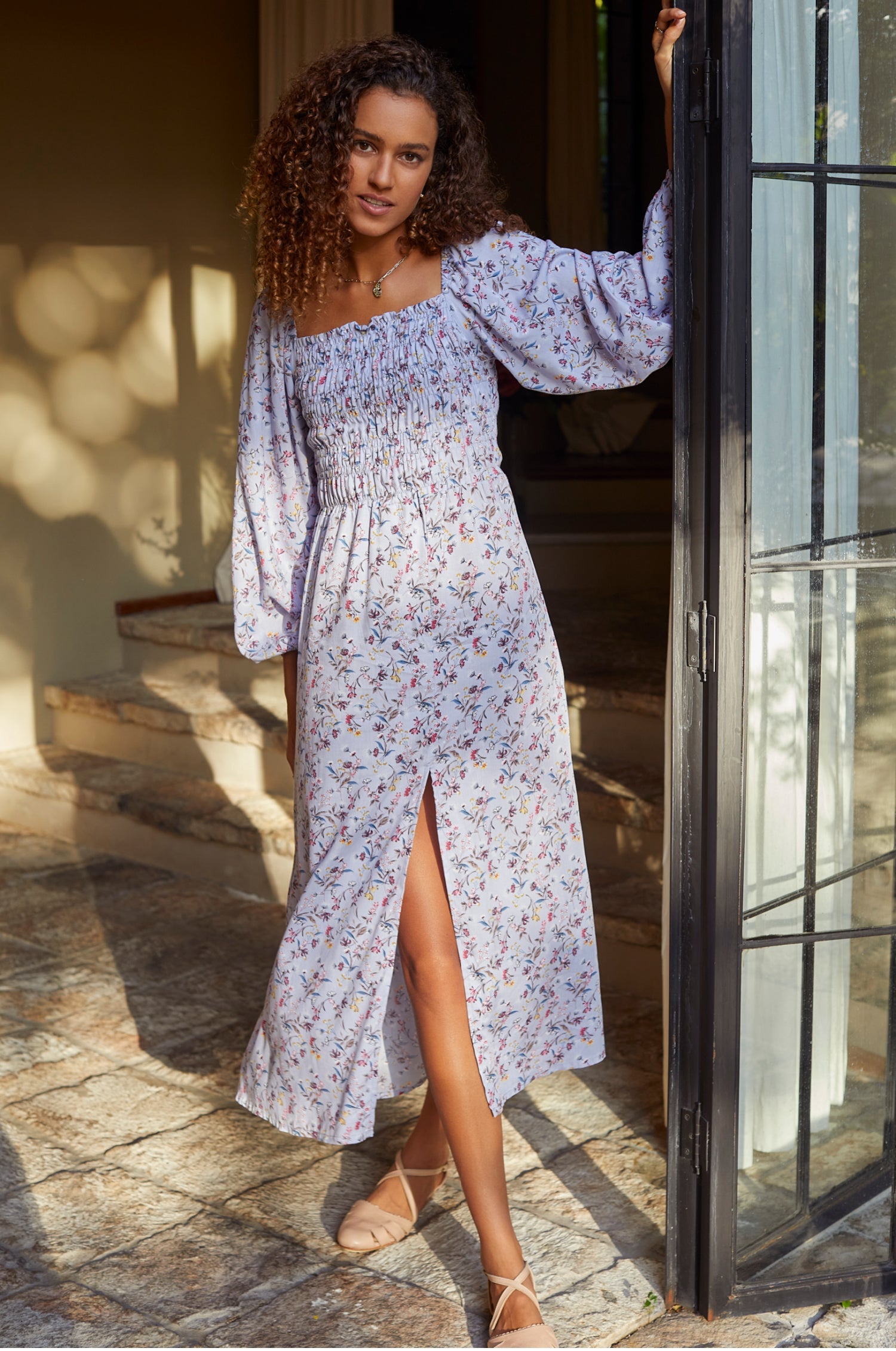 Woman in a floral dress standing by a door with sunlight filtering through.