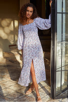 Woman in a floral dress standing by a door with sunlight filtering through.