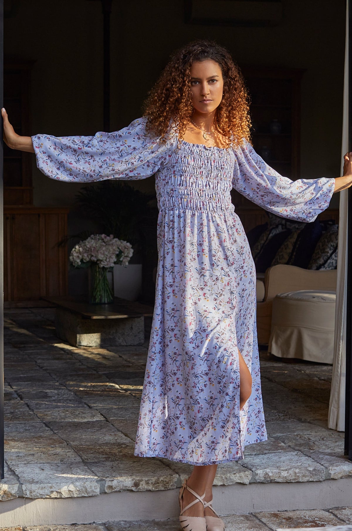 Woman in a long, floral dress standing in a sunlit room with flowers in the background.