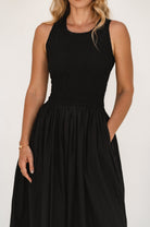 Woman wearing a black sleeveless dress with shirred bodice and hand in pocket against a white background. 