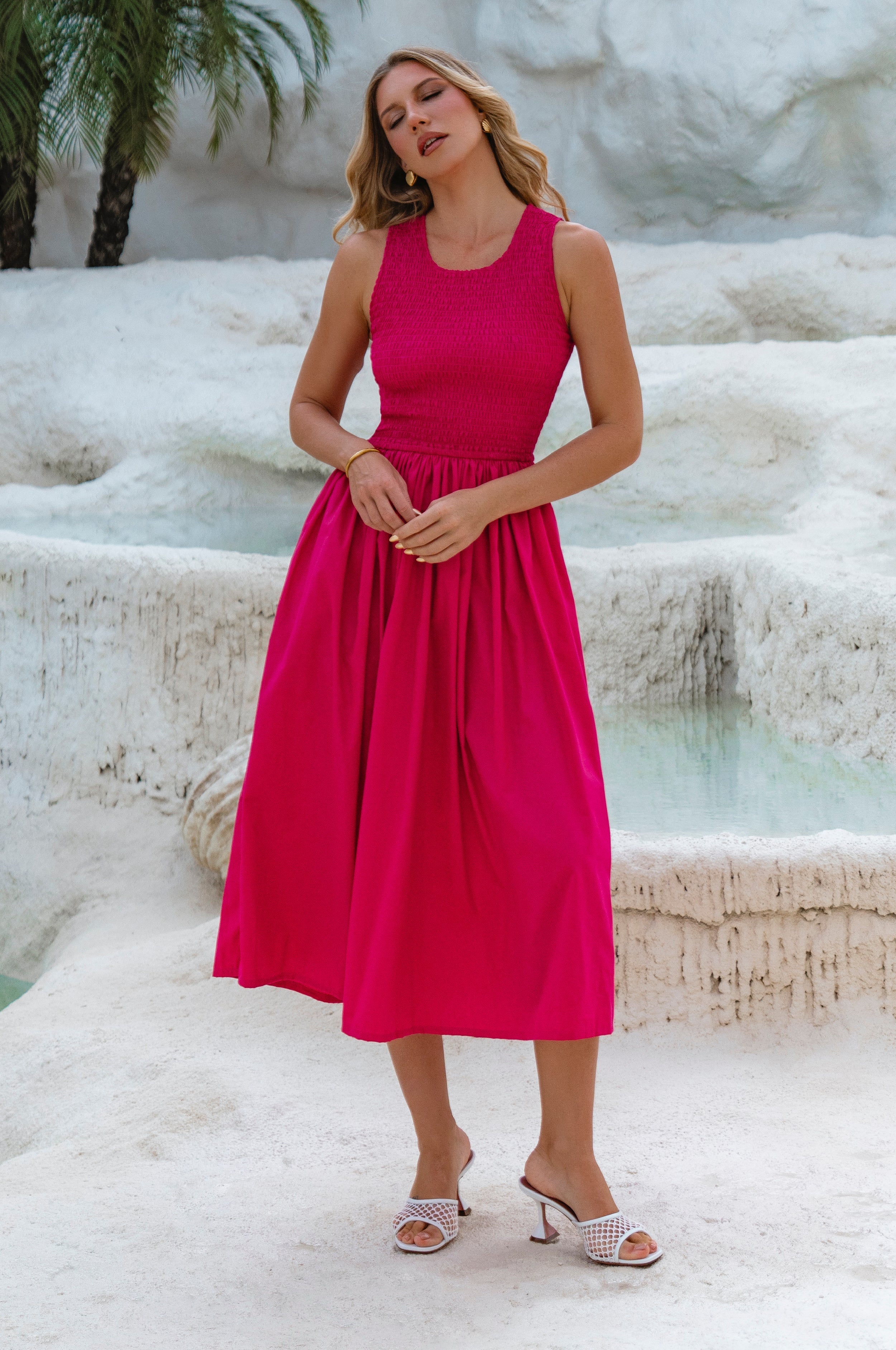 Woman in a bright pink cotton sleeveless midi dress standing in front of a natural water feature