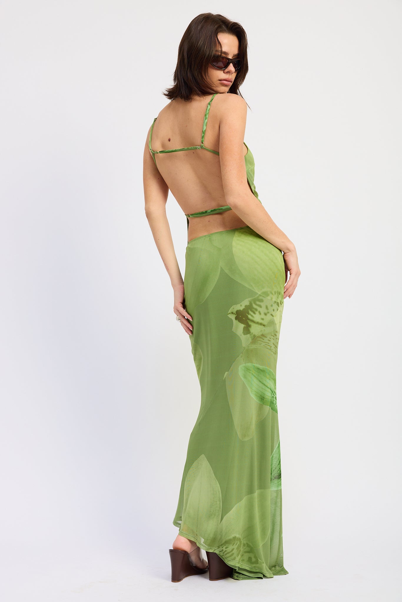 Woman wearing a green orchid print floral maxi skirt and matching open back cami top with slim straps sleeveless top with a white background back view