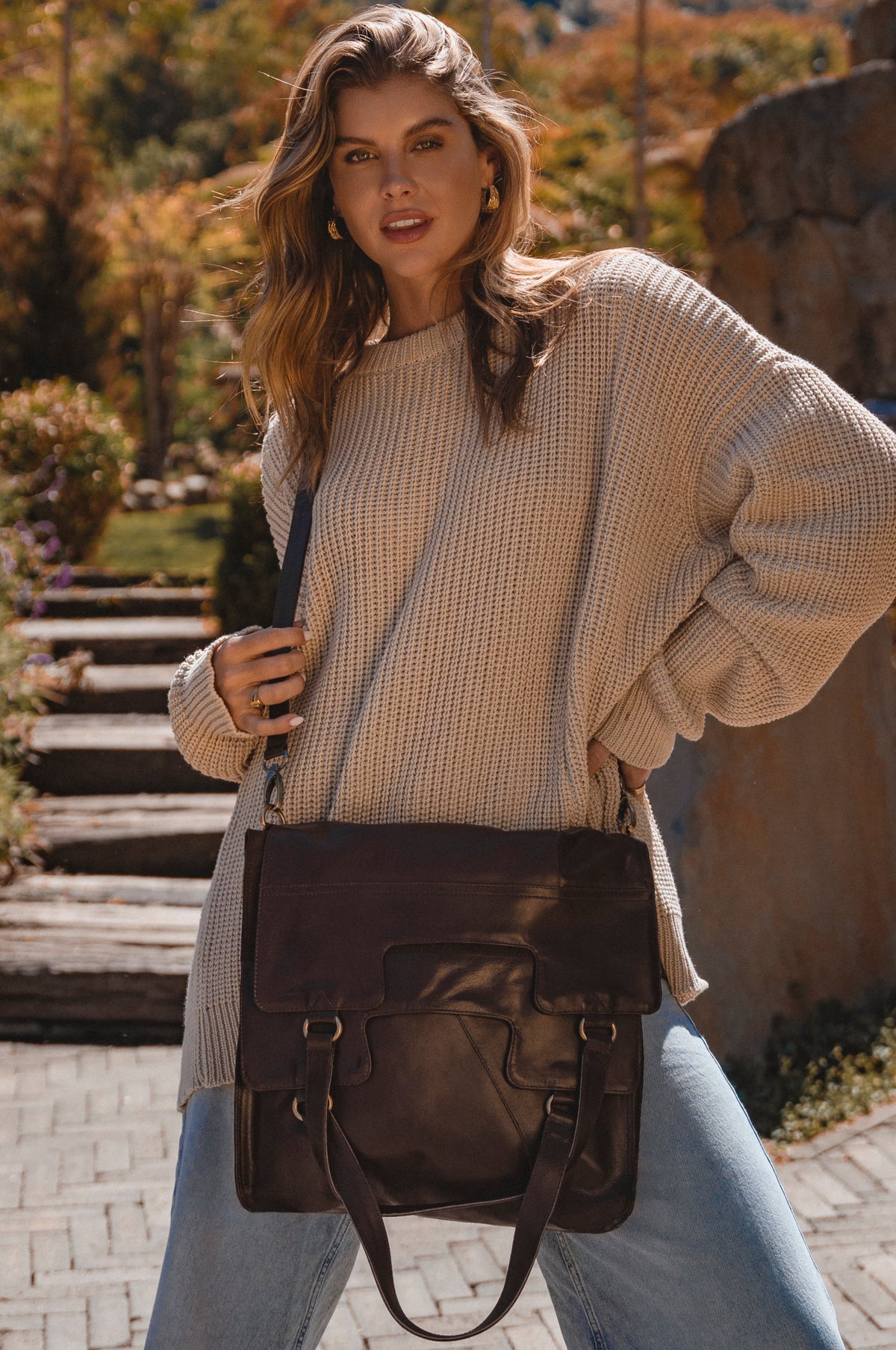 Woman holding a brown leather bag outdoors with trees and steps in the background