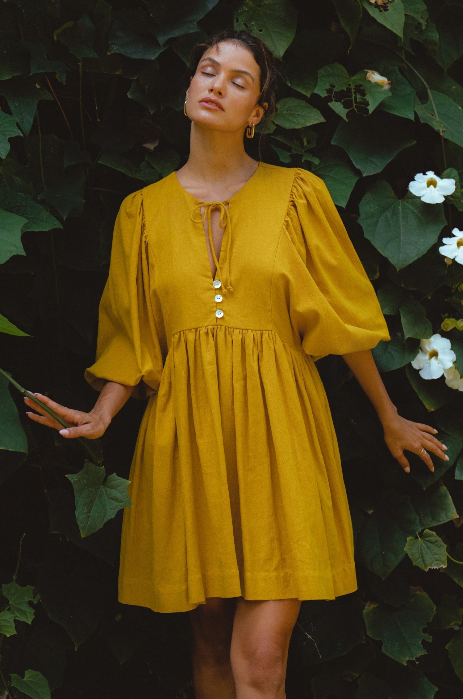 Woman wearing mustard yellow colored linen mini dress or lightweight crinkle linen in a long-sleeve babydoll silhouette against a vine covered wall, front view
