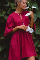 Woman wearing wine colored linen mini dress or lightweight crinkle linen in a long-sleeve babydoll silhouette against a vine covered wall, front view