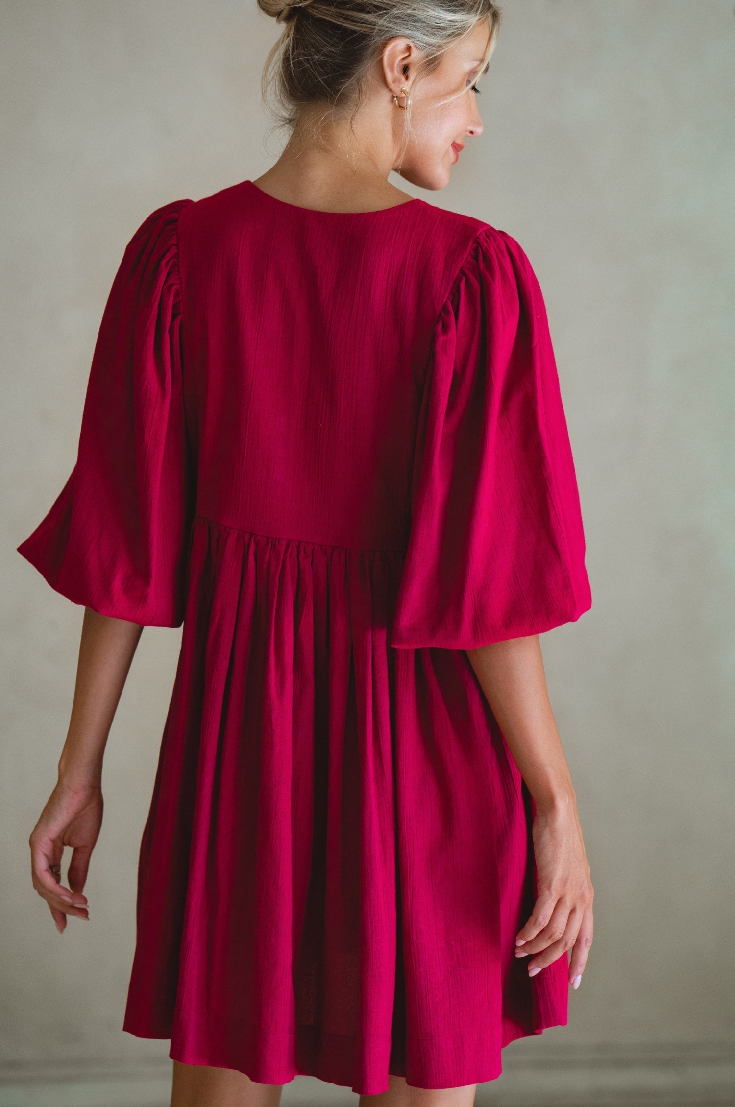 Woman wearing wine colored linen mini dress or lightweight crinkle linen in a long-sleeve babydoll silhouette against a neutral background, back view