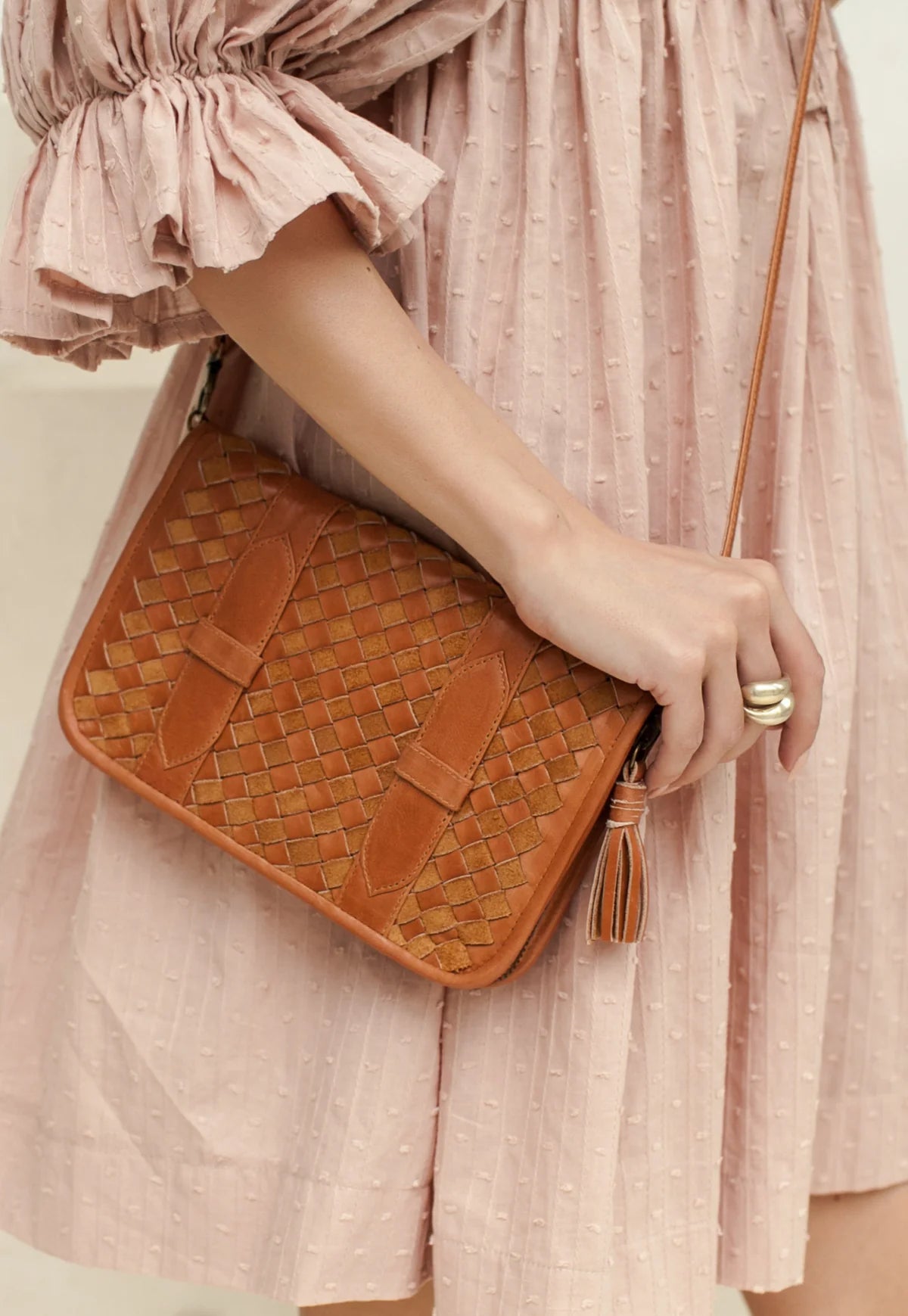 A woman in a textured light pink dress holds a tan woven leather crossbody bag with a tassel zipper pull.