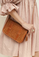 A woman in a textured light pink dress holds a tan woven leather crossbody bag with a tassel zipper pull.