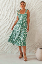Woman wearing a green patterned square neckline and button-down fit-and-flare midi dress with adjustable tie-up straps and a smocked back against a neutral background, front view