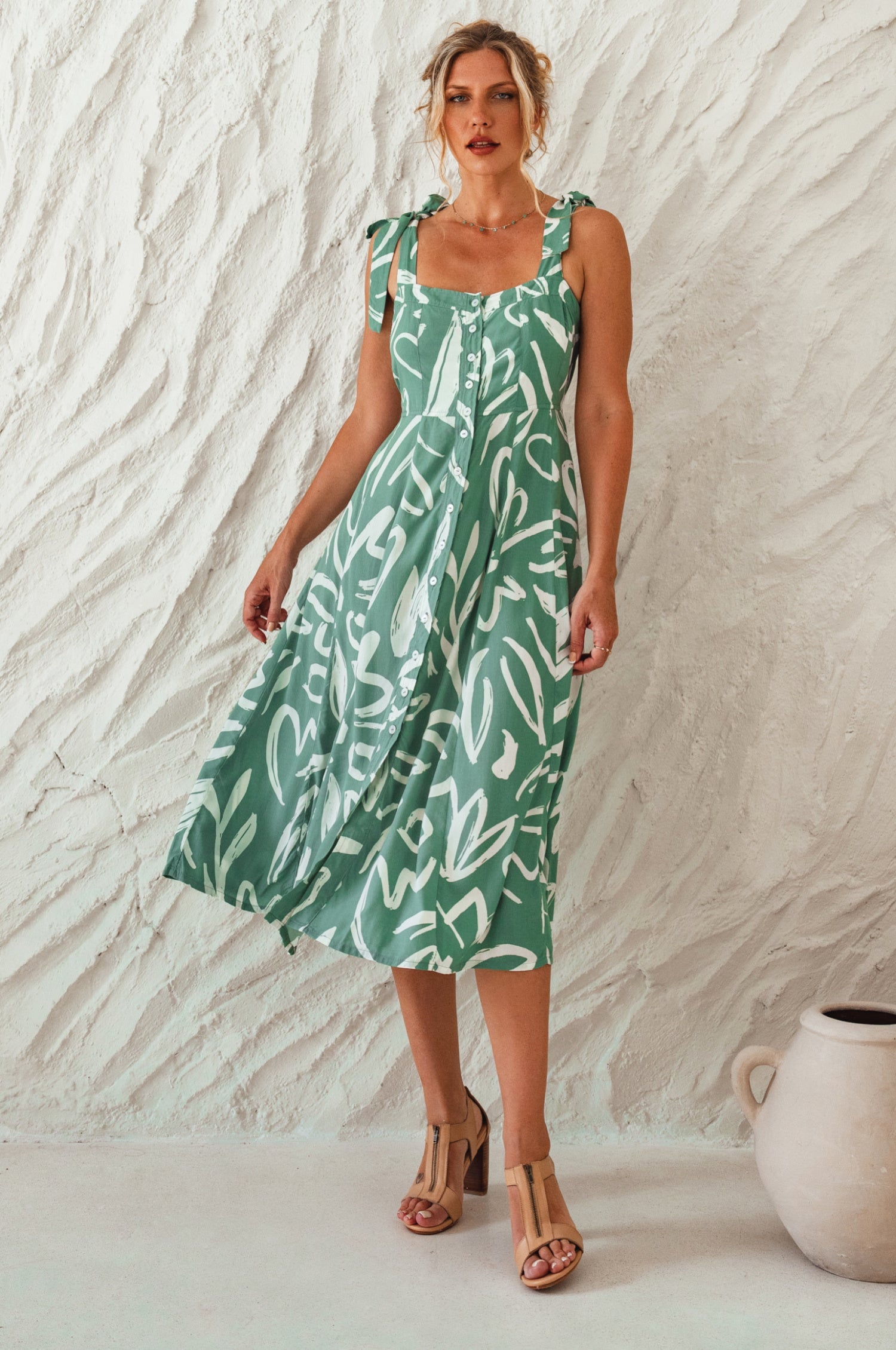 Woman wearing a green patterned square neckline and button-down fit-and-flare midi dress with adjustable tie-up straps and a smocked back against a neutral background, front view