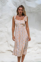 Woman wearing a cream floral square neckline and button-down fit-and-flare midi dress with adjustable tie-up straps and a smocked back against a neutral background, front view