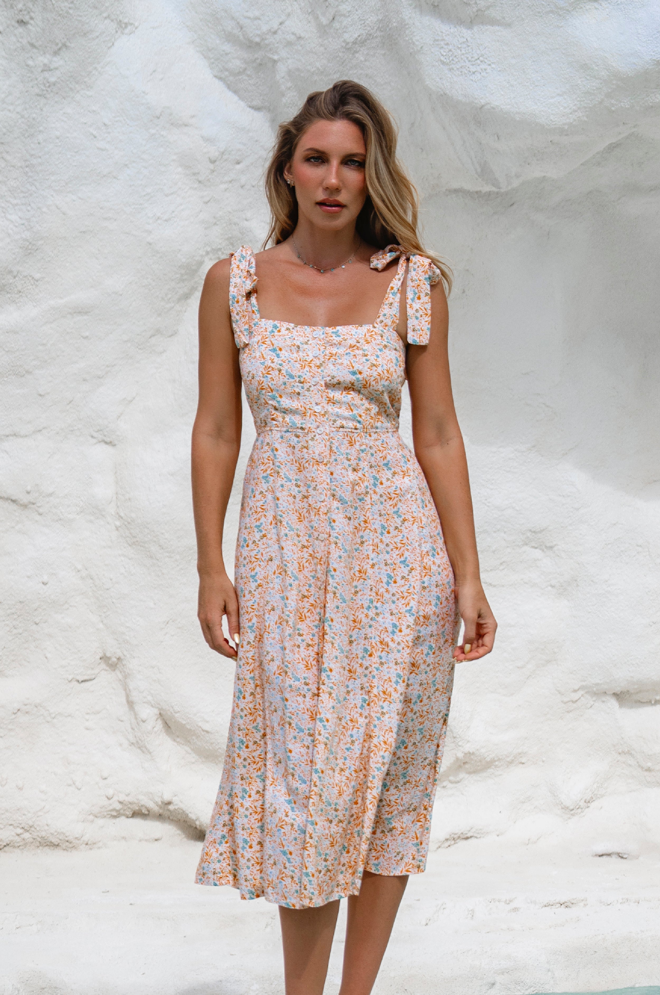 Woman wearing a cream floral square neckline and button-down fit-and-flare midi dress with adjustable tie-up straps and a smocked back against a neutral background, front view