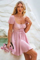 Woman wearing pink mini dress with corset-style bodice and matching ties on the front with elasticated sleeves worn on the shoulder holding flowers against a white textured background
