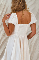 Woman wearing ivory mini dress with corset-style bodice and matching ties on the front with elasticated sleeves worn on the shoulder with white background, back view.