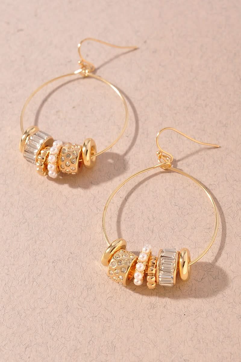 Gold hoop earrings with decorative bead clusters featuring clear crystals, white pearls, and textured gold discs, displayed on a textured beige background.
