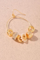 Gold hoop earring with decorative charms, including a textured gold bead, a clear crystal bead, and a pearl bead, displayed on a textured beige background.