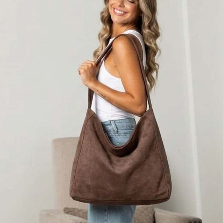 A woman wearing a white tank top and light blue jeans carries a large dark brown suede hobo shoulder bag in a bright indoor setting.