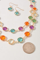 A gold-tone necklace and matching drop earrings with round, colorful gemstone bezel settings, displayed on a light background with a US quarter coin for scale.