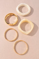 Five bangle bracelets arranged on a warm beige background: three slim gold-tone bands and two chunkier marbled beige bands with organic shapes.