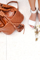 A camel woven leather bucket bag with braided straps and a tassel detail, styled on a light beigh=e stone surface next to a model's feet wearing white woven sandals.