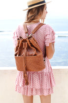 A woman wearing a pink and white polka dot dress and straw hat, seen from behind, carrying a camel woven leather bucket backpack with a tassel drawstring and braided straps against a beach background.