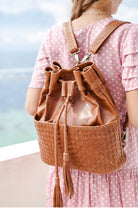 A woman wearing a pink polka-dot dress carries a camel leather drawstring backpack with a woven front pocket, photographed outdoors with a blurred sky and water background.