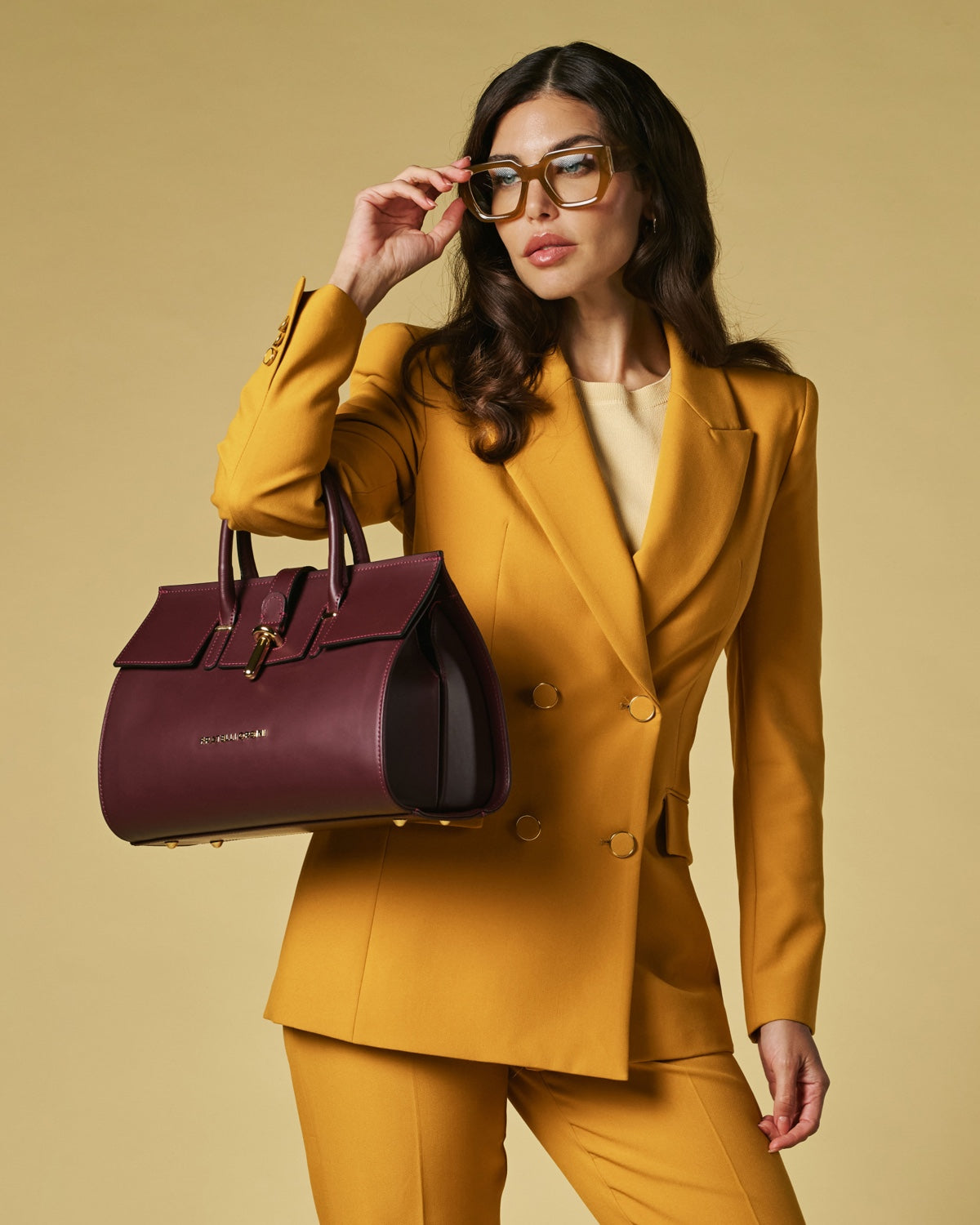 Woman in a mustard yellow suit holding a maroon handbag against a beige background