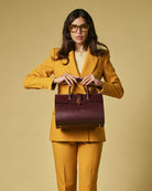 Woman in a mustard yellow suit holding a maroon handbag against a beige background