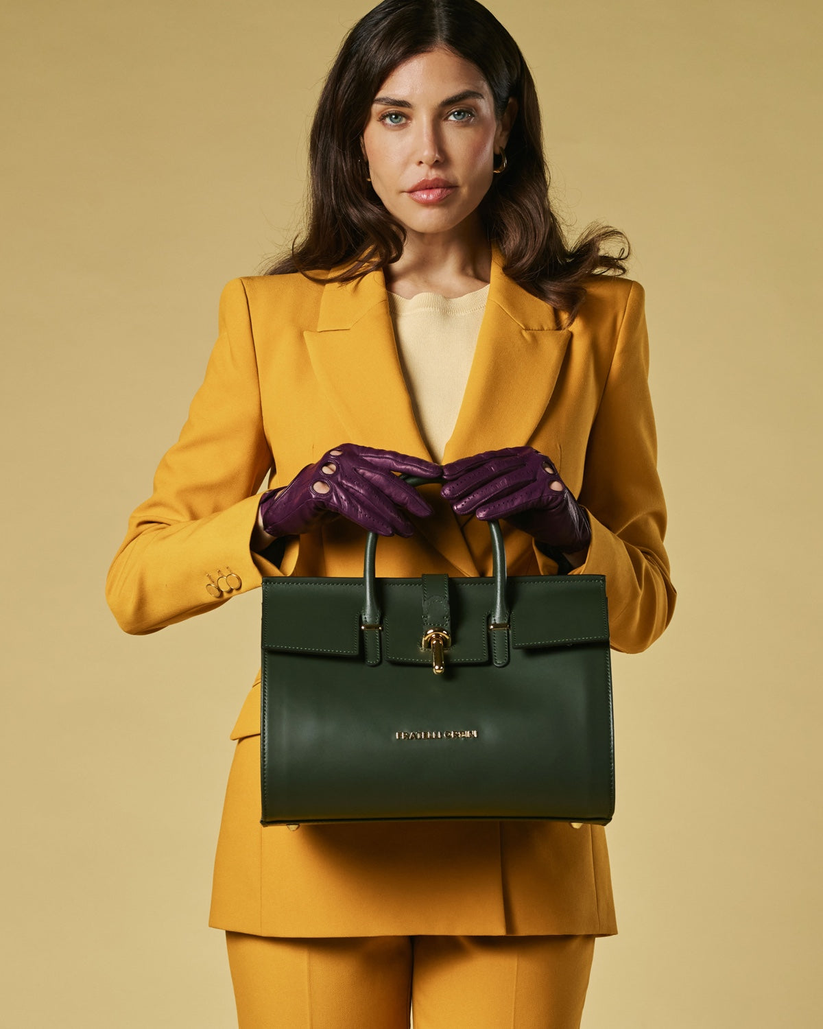 Woman in mustard suit holding a green handbag against a beige background