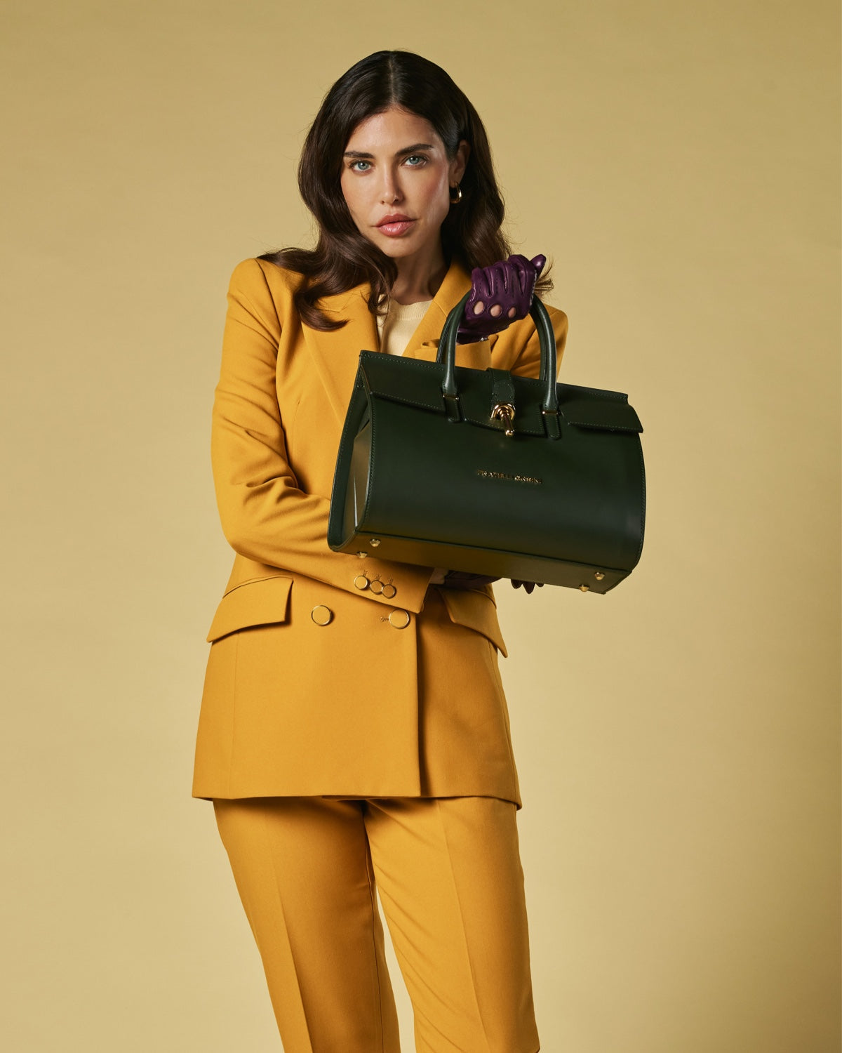 Woman in a mustard yellow suit holding a green leather handbag against a beige background