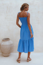 Woman wearing a blue linen tiered midi dress with wooden bead adorned spaghetti straps and smocked bodice standing against a plain background, back view