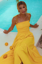 Woman wearing a bright yellow linen tiered midi dress with wooden bead adorned spaghetti straps and smocked bodice  sitting by a pool