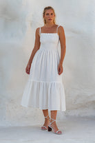 Woman wearing an off-white linen tiered midi dress with wooden bead adorned spaghetti straps and smocked bodice against a plain white background