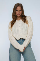 Woman wearing a cream knitted sweater and blue jeans against a plain background