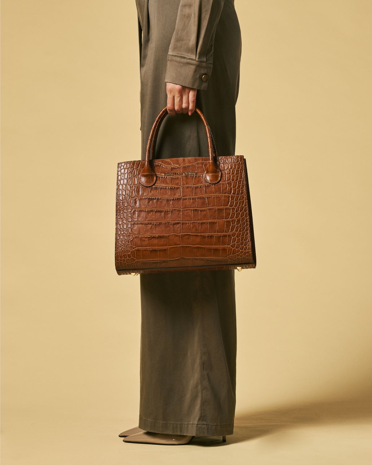 Brown leather handbag held by a person wearing a green outfit against a beige background