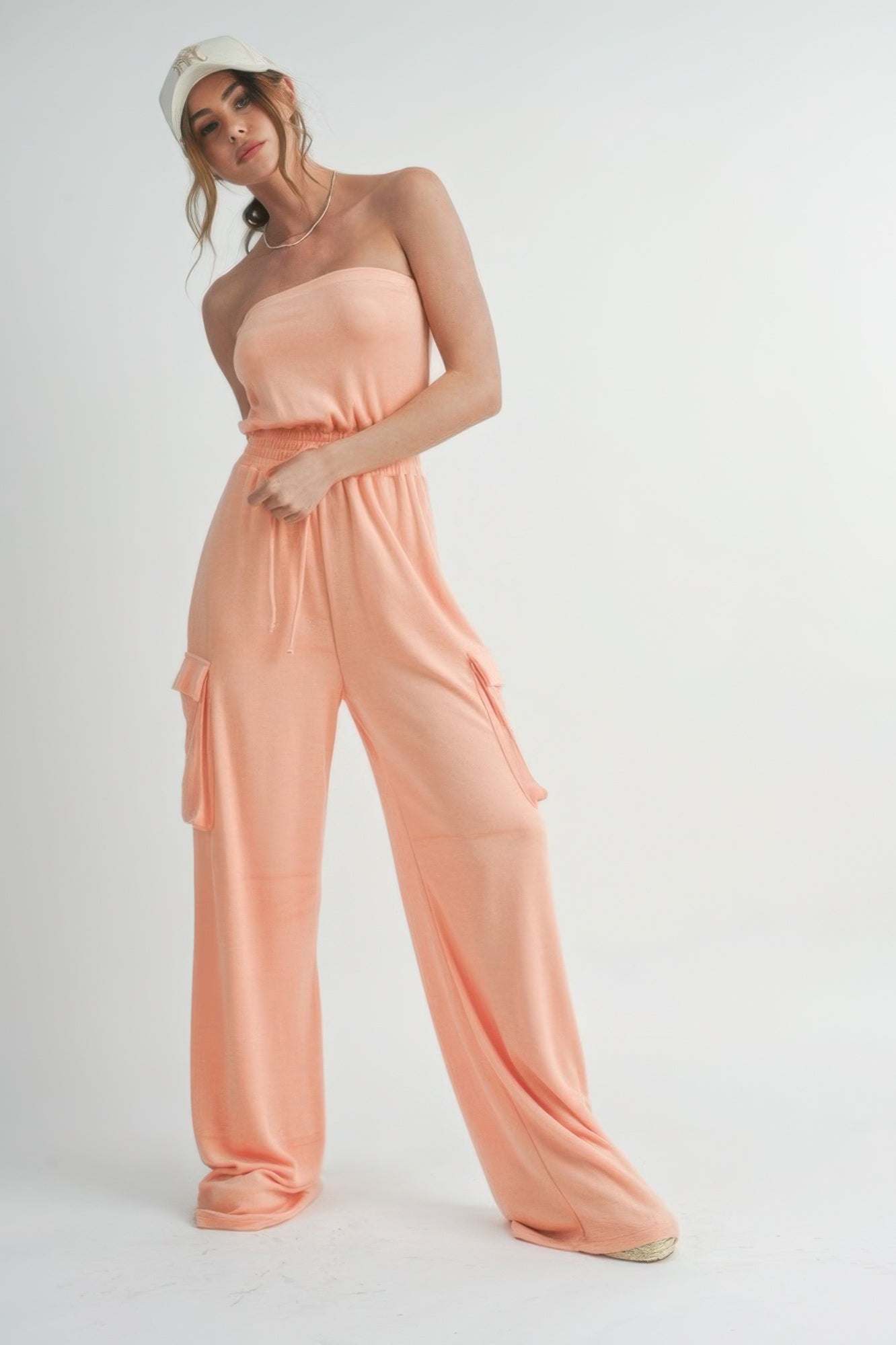 Front view of model wearing peach strapless cargo jumpsuit with smocked waist and wide legs styled with baseball cap.