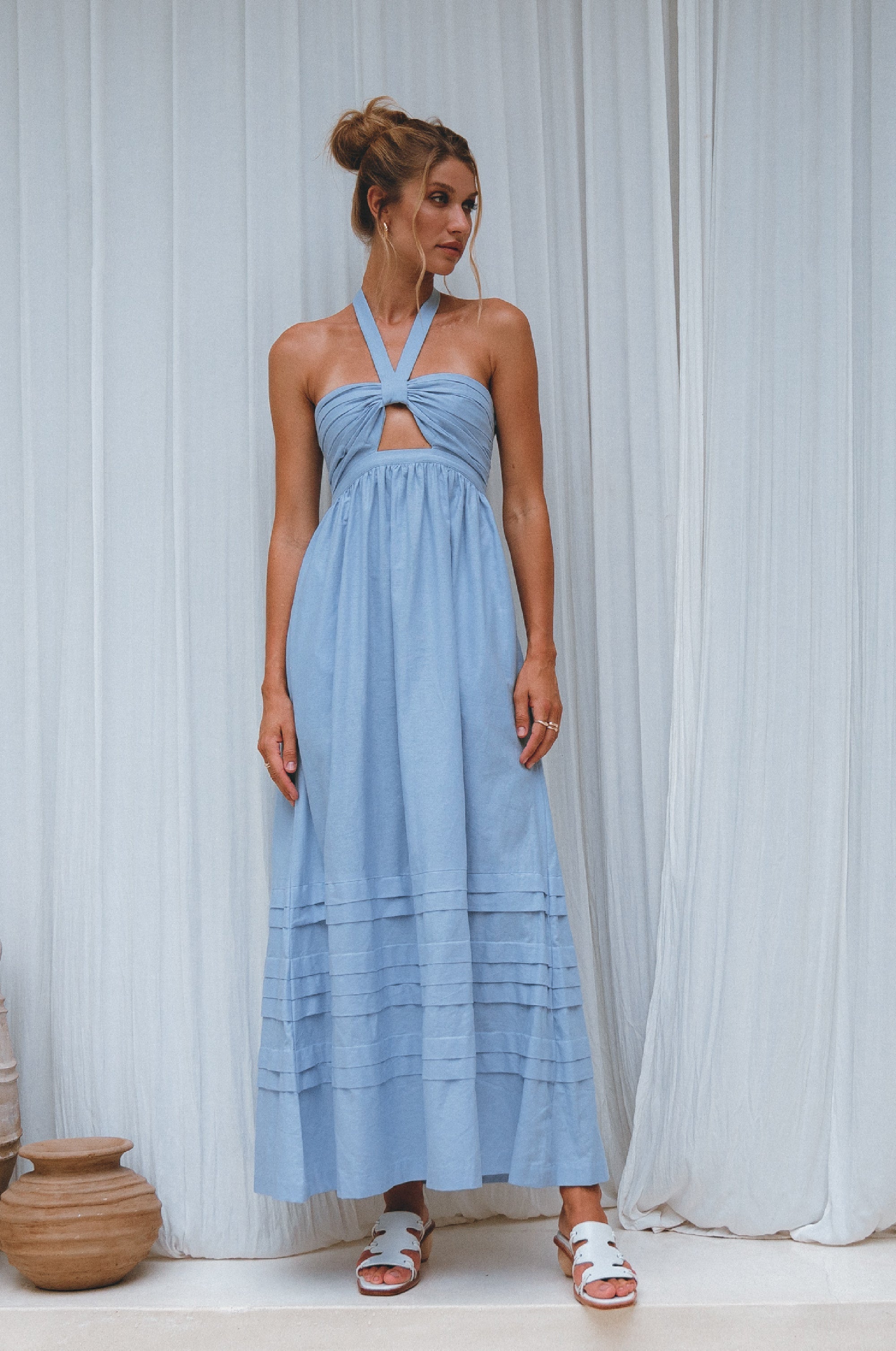 Woman wearing a light blue linen maxi dress with halter neck and front cut out standing against a white curtain.